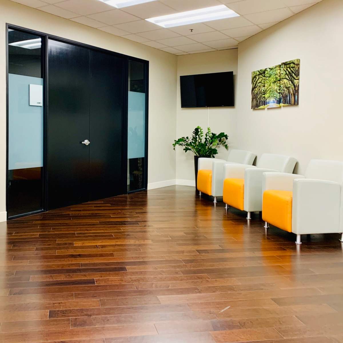 A modern waiting area with orange and white chairs, a black glass door, and a wall-mounted TV, set against a wooden floor.