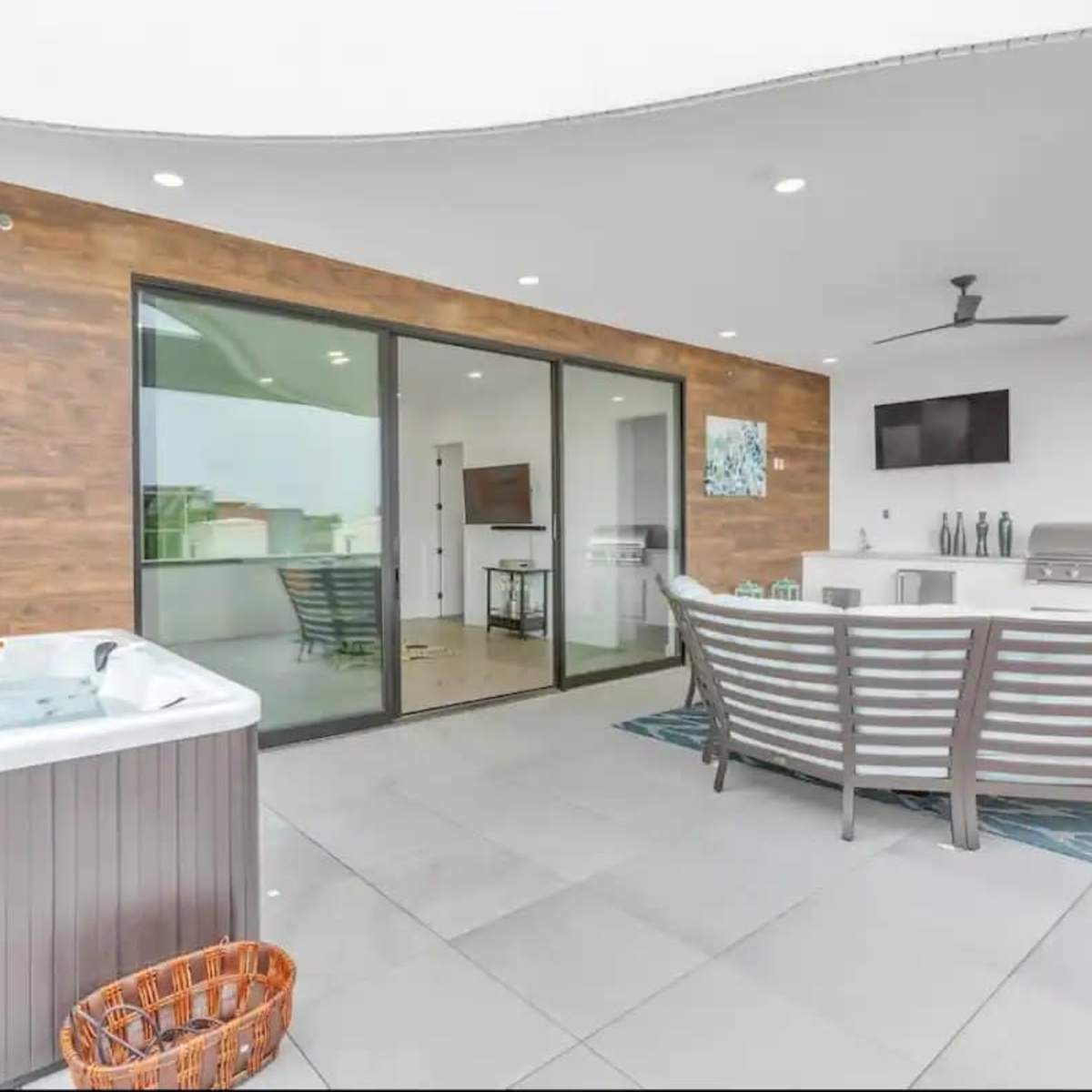 A modern outdoor space featuring a hot tub, seating area, and a grill station with sliding glass doors leading to an interior room.