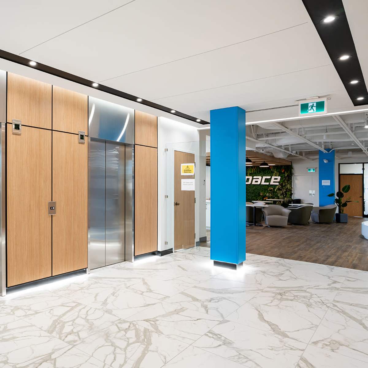 A modern lobby area with wooden elevator doors, a sleek metal elevator, and a blue wall featuring a sign, along with a marble floor and seating area in the background.