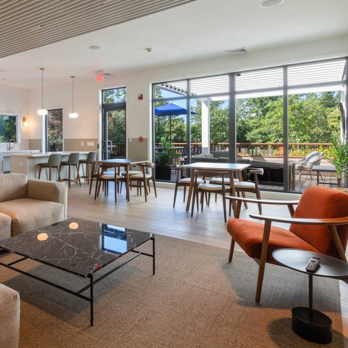 A modern lounge area with a beige sectional sofa, a coffee table, and dining tables positioned near large windows that overlook an outdoor space.