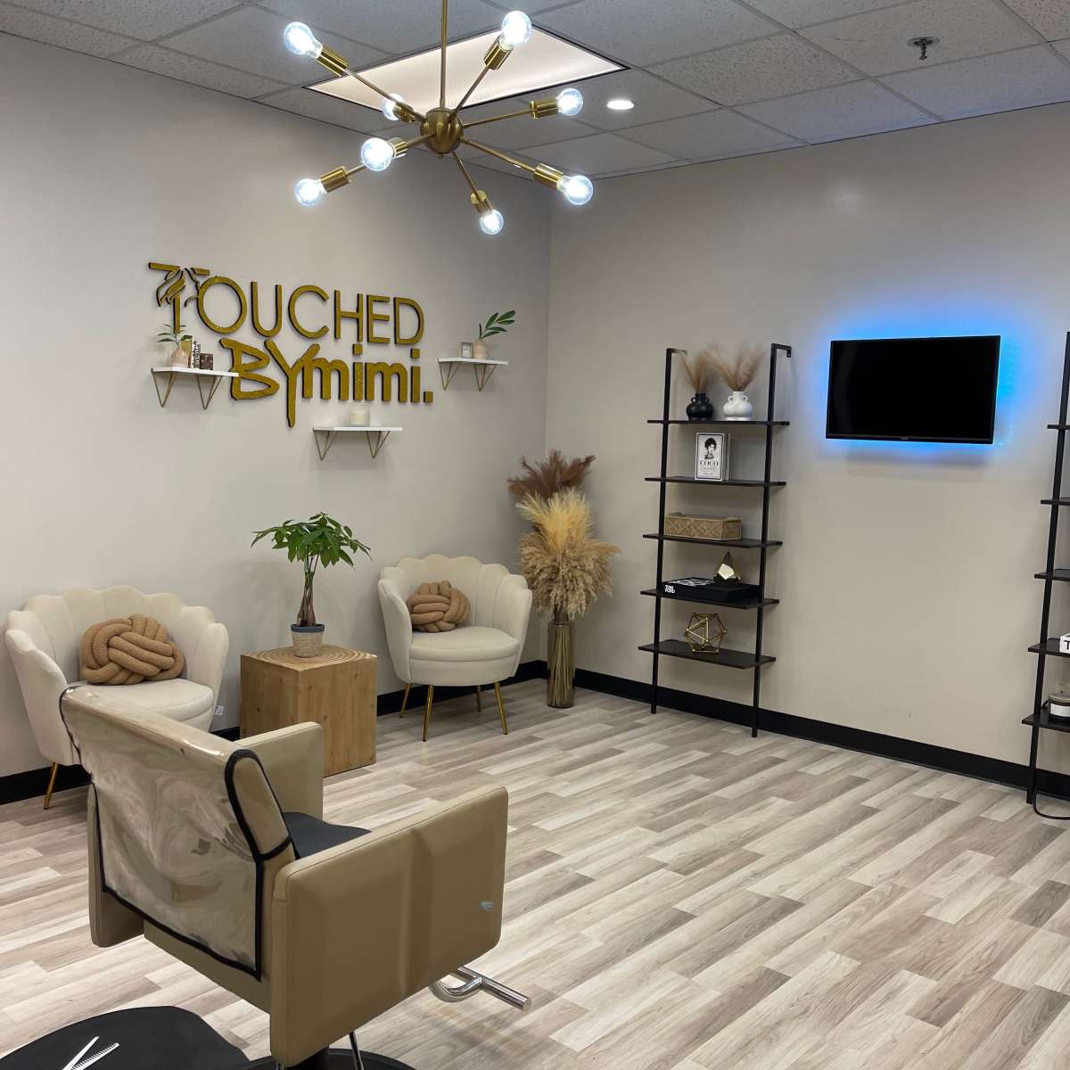 A salon interior with two white chairs, a wooden table, decorative plants, and a wall sign that reads "Touched By Mimi" in gold lettering.