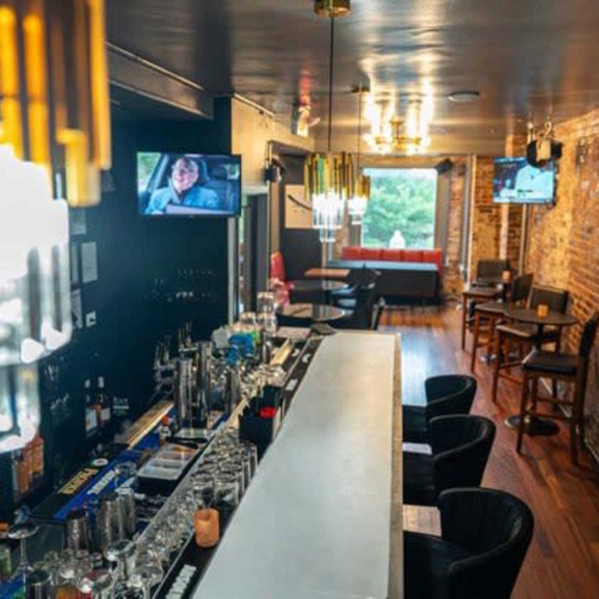 A modern bar scene features a long white countertop with various drinks positioned on it, surrounded by black chairs, and two televisions displaying content on the walls of a brick interior.