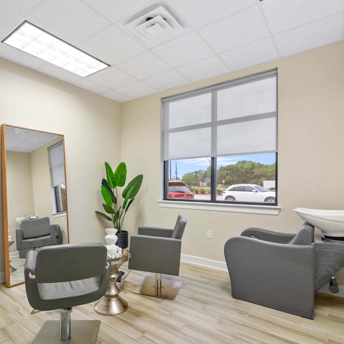 A modern salon interior with two gray chairs, a large mirror, a potted plant, and a hair wash station beside a window.