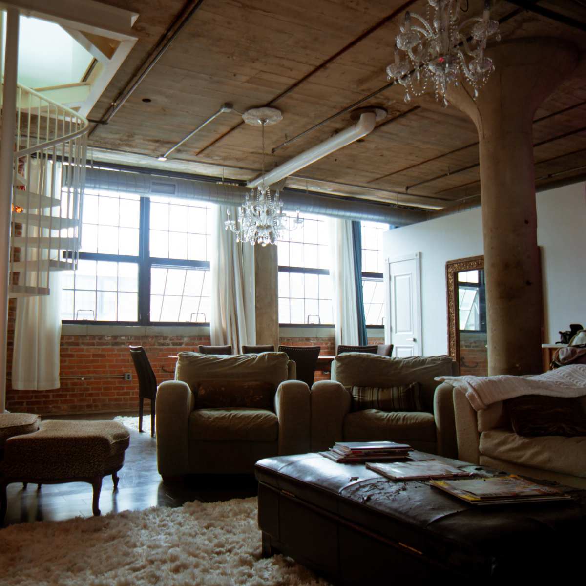 A modern loft interior featuring a spiral staircase, large windows, and a mix of furniture including armchairs and a coffee table on a plush rug.