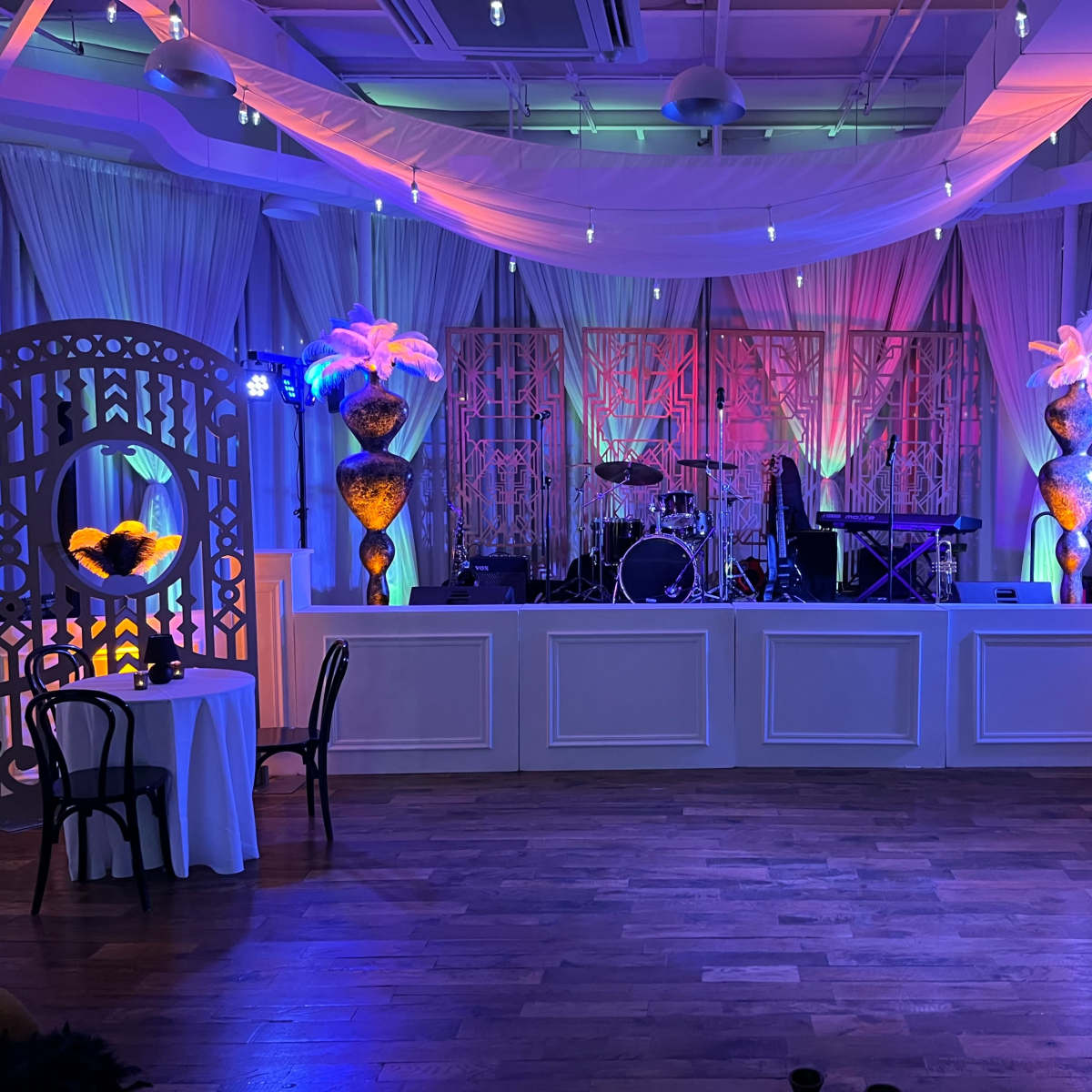 A decorated event space featuring a stage with musical instruments, illuminated by colored lights, and adorned with floral arrangements on either side.
