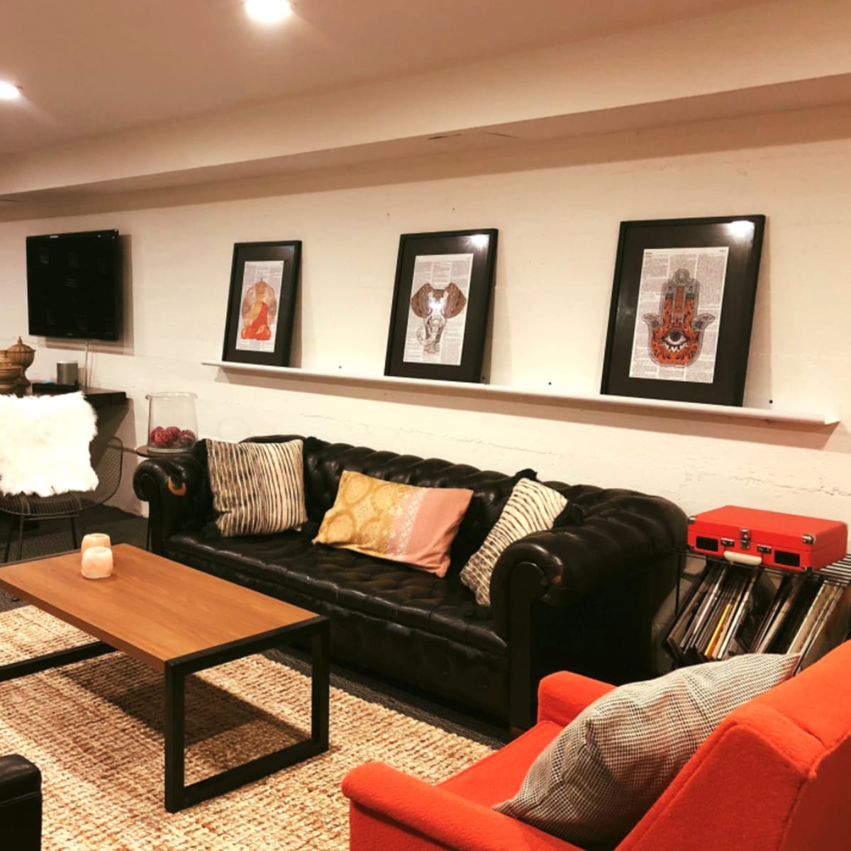 A cozy basement lounge area with a black leather sofa, an orange chair, a wooden coffee table, and framed artwork on the wall.