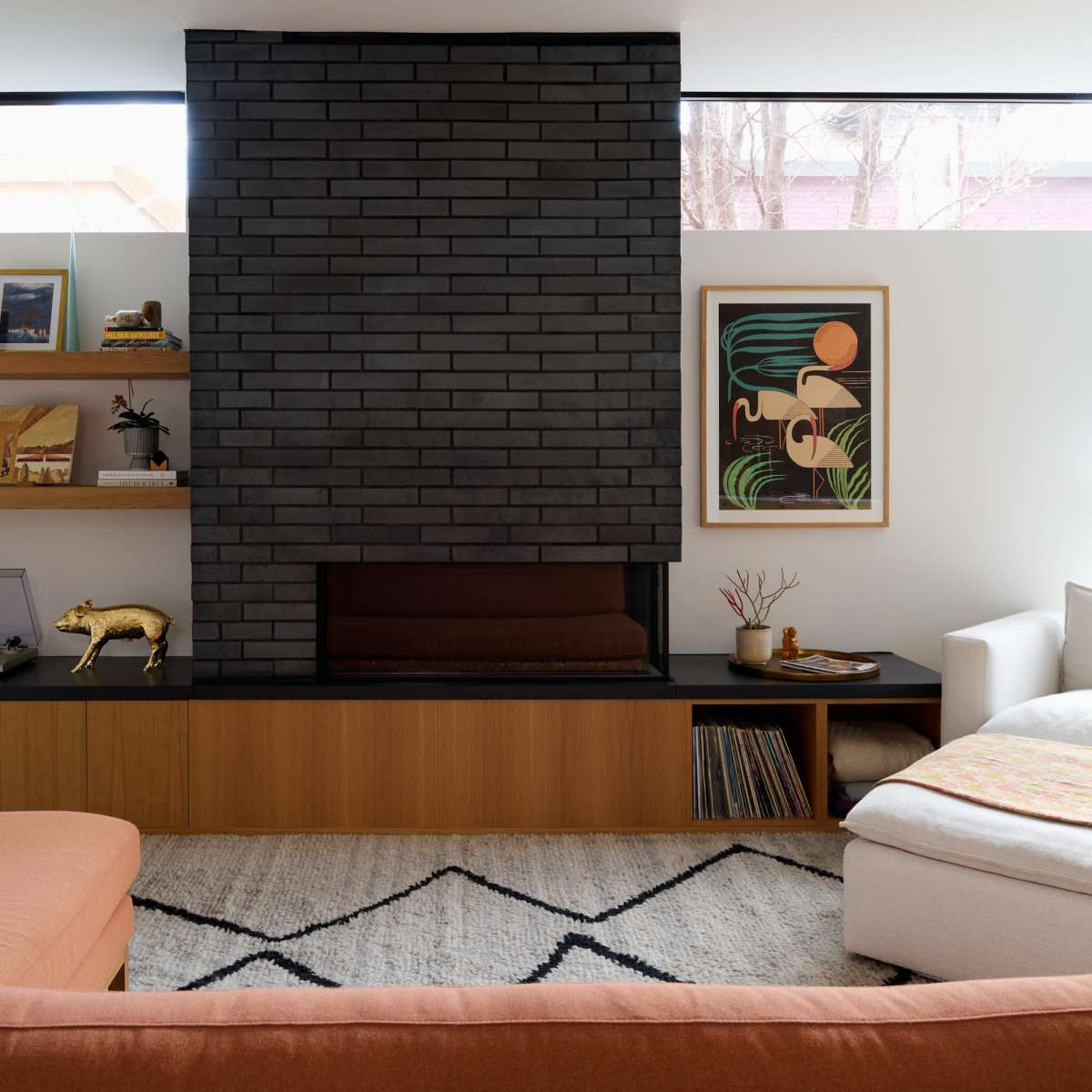A modern living room featuring a dark brick fireplace, wooden shelves with decorative items, and a light-colored sofa paired with an orange sectional couch.