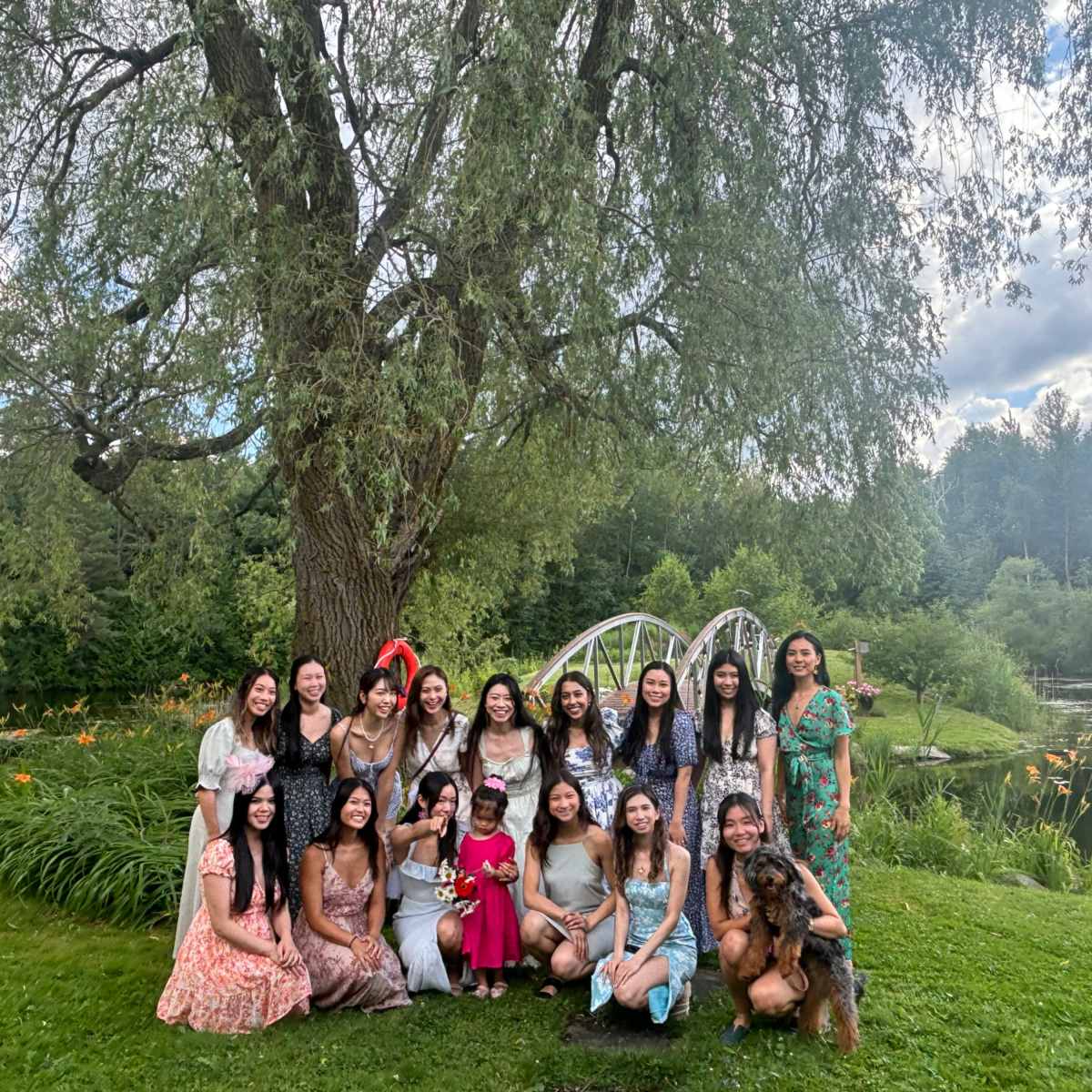 A group of women poses together in a lush garden near a pond with a decorative bridge in the background.