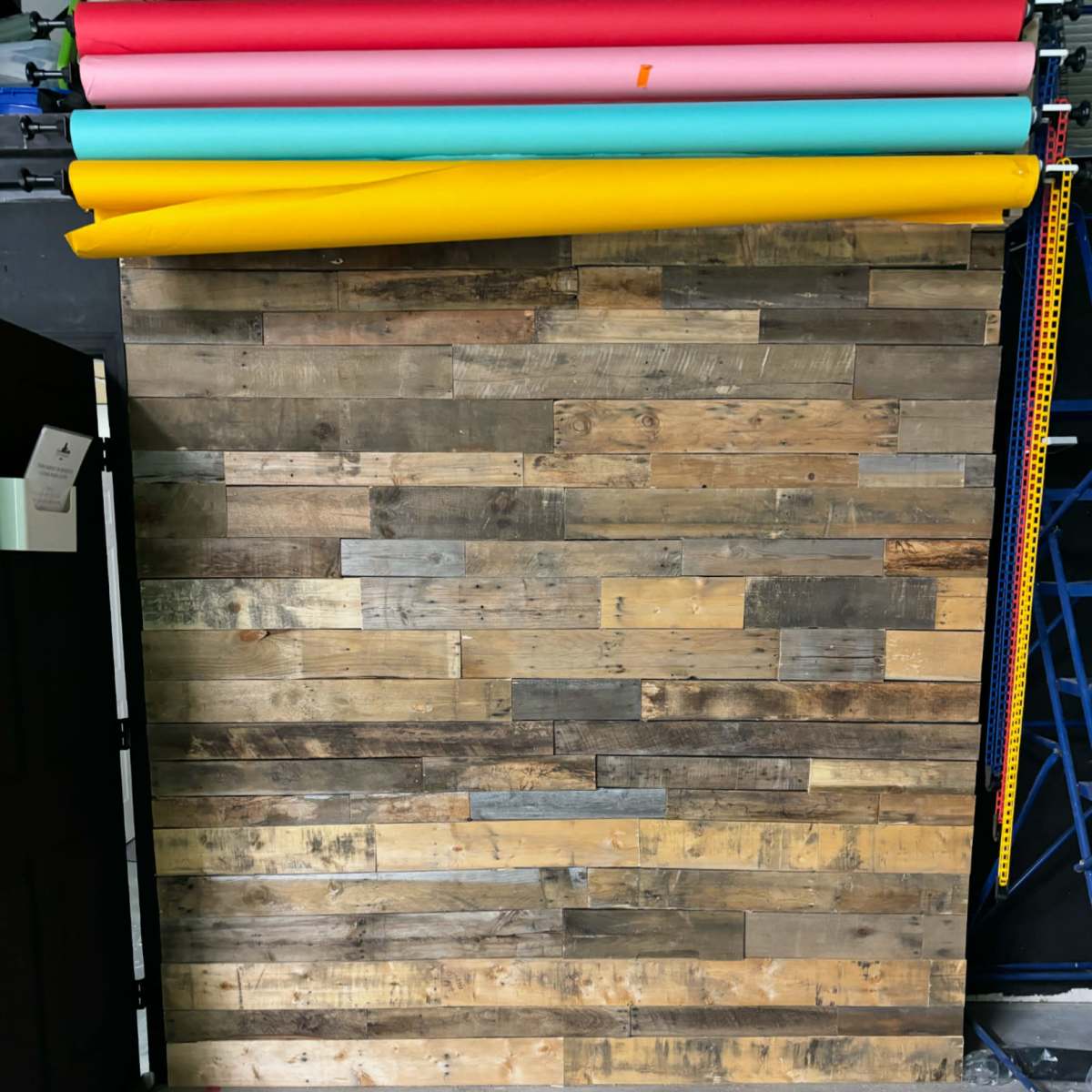 A wooden wall made up of variously colored planks is placed in front of a rack holding rolls of colored backdrop paper.