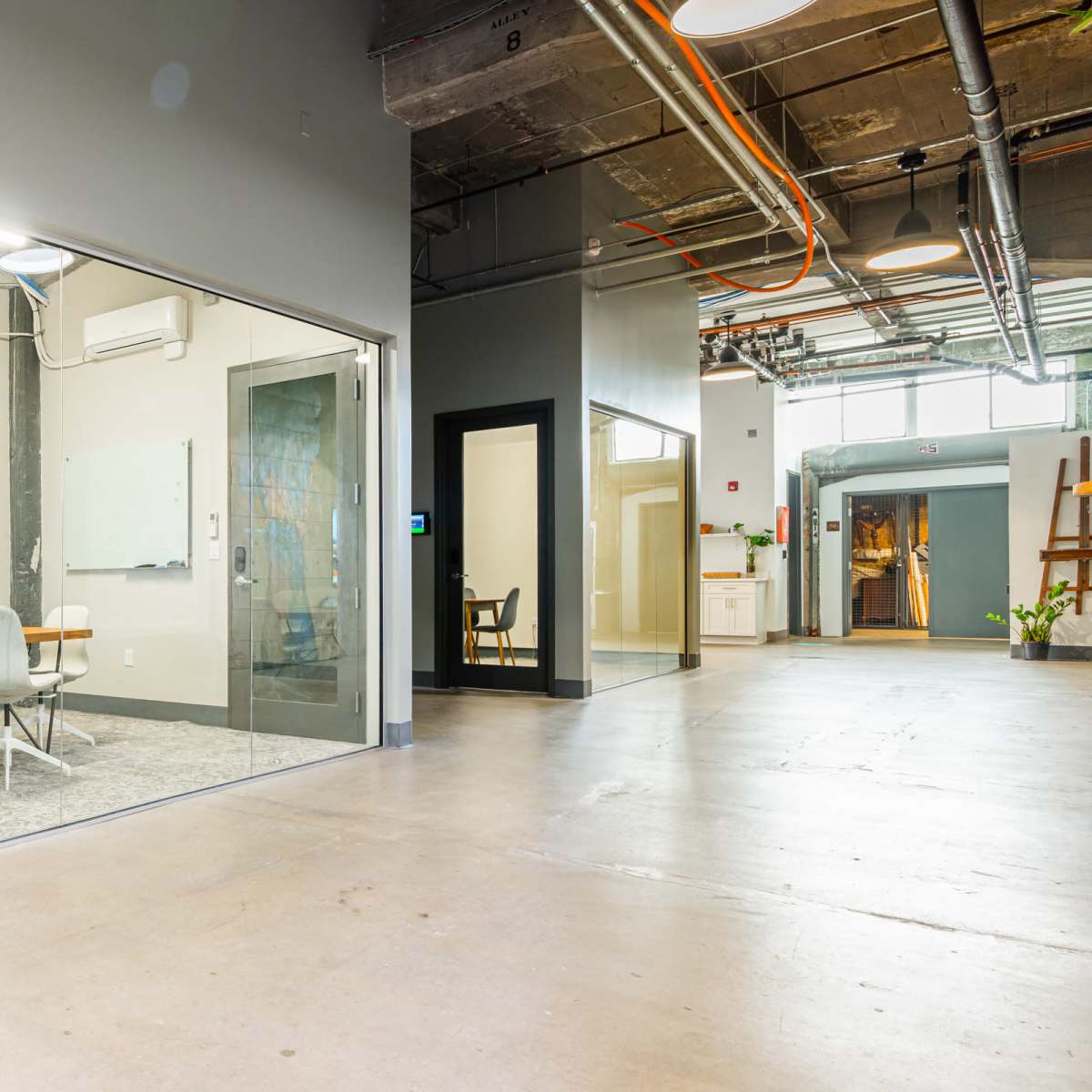 A modern interior space with a glass-walled conference room on the left and an office door on the right, all situated within a large open area.
