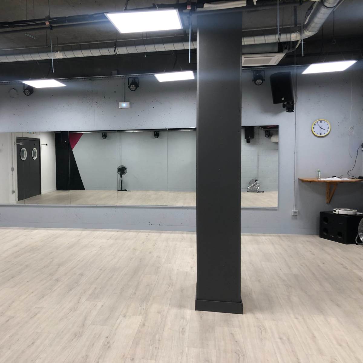 A spacious, empty studio with wooden flooring, mirrored walls, and a clock on one wall.
