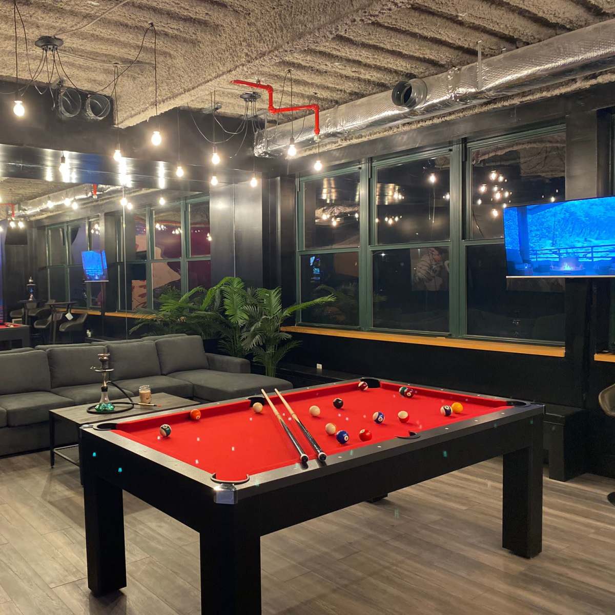A game room featuring a red pool table, a large gray sectional sofa, and wall-mounted televisions, all illuminated by pendant lights.