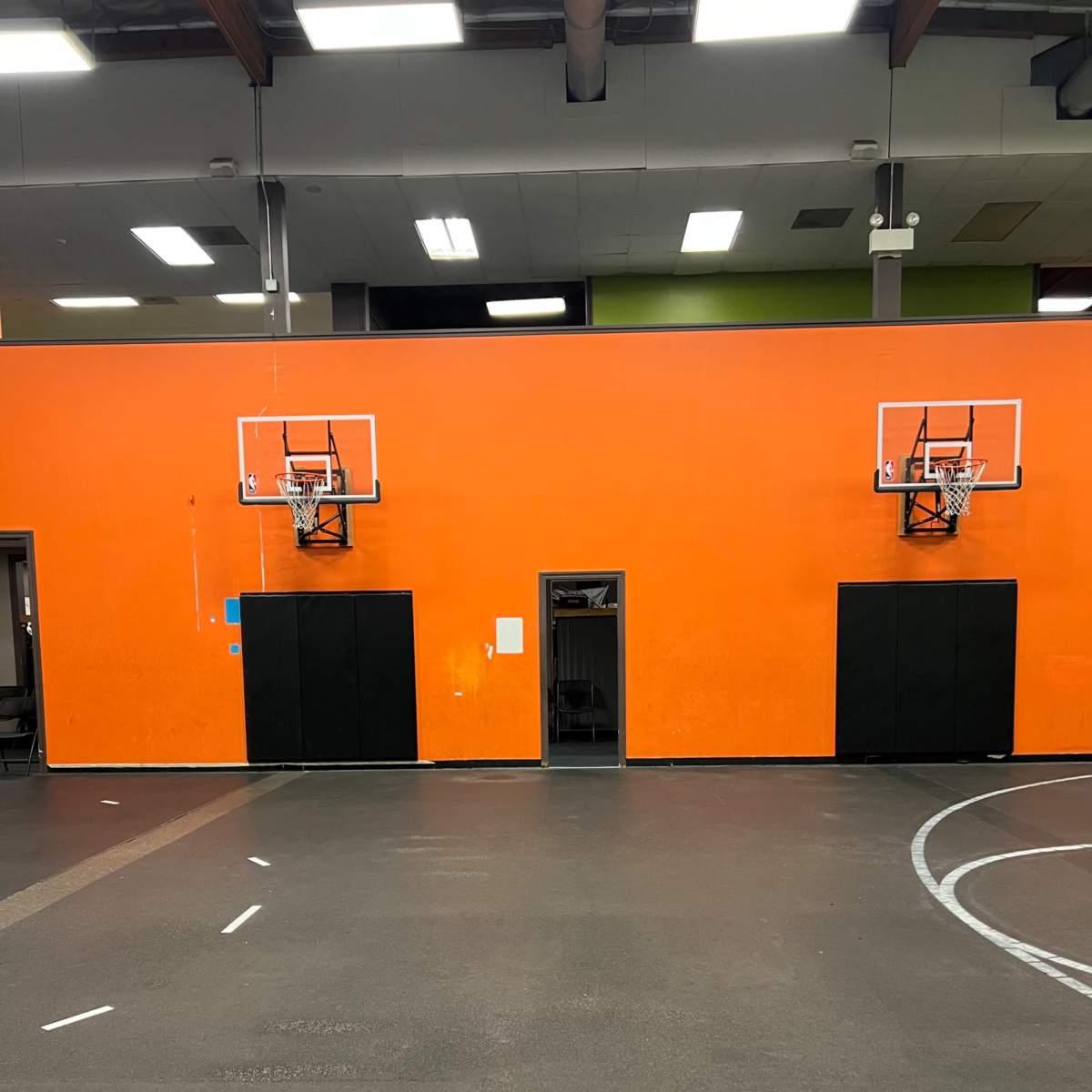 A basketball court with two hoops mounted on an orange wall, flanked by black cabinets.