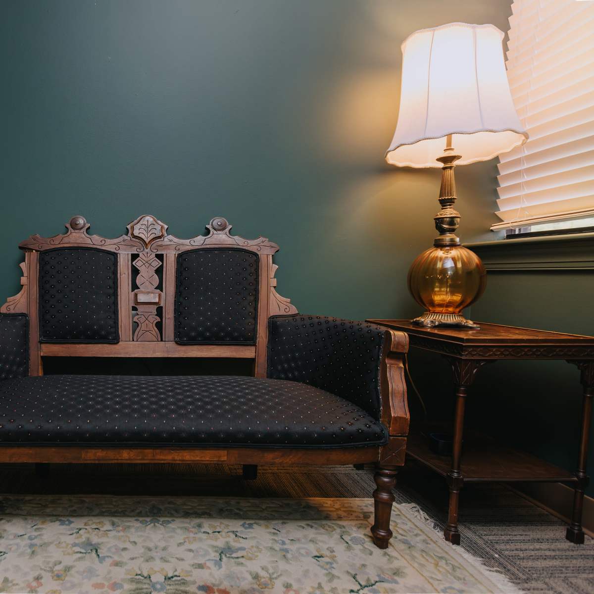A dark wooden loveseat with a patterned cushion, positioned against a green wall next to a lamp and a small side table.