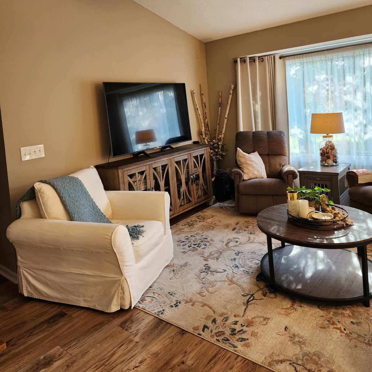 A cozy living room featuring a television mounted on the wall, a circular coffee table in the center, and a beige sofa with a blue throw pillow.