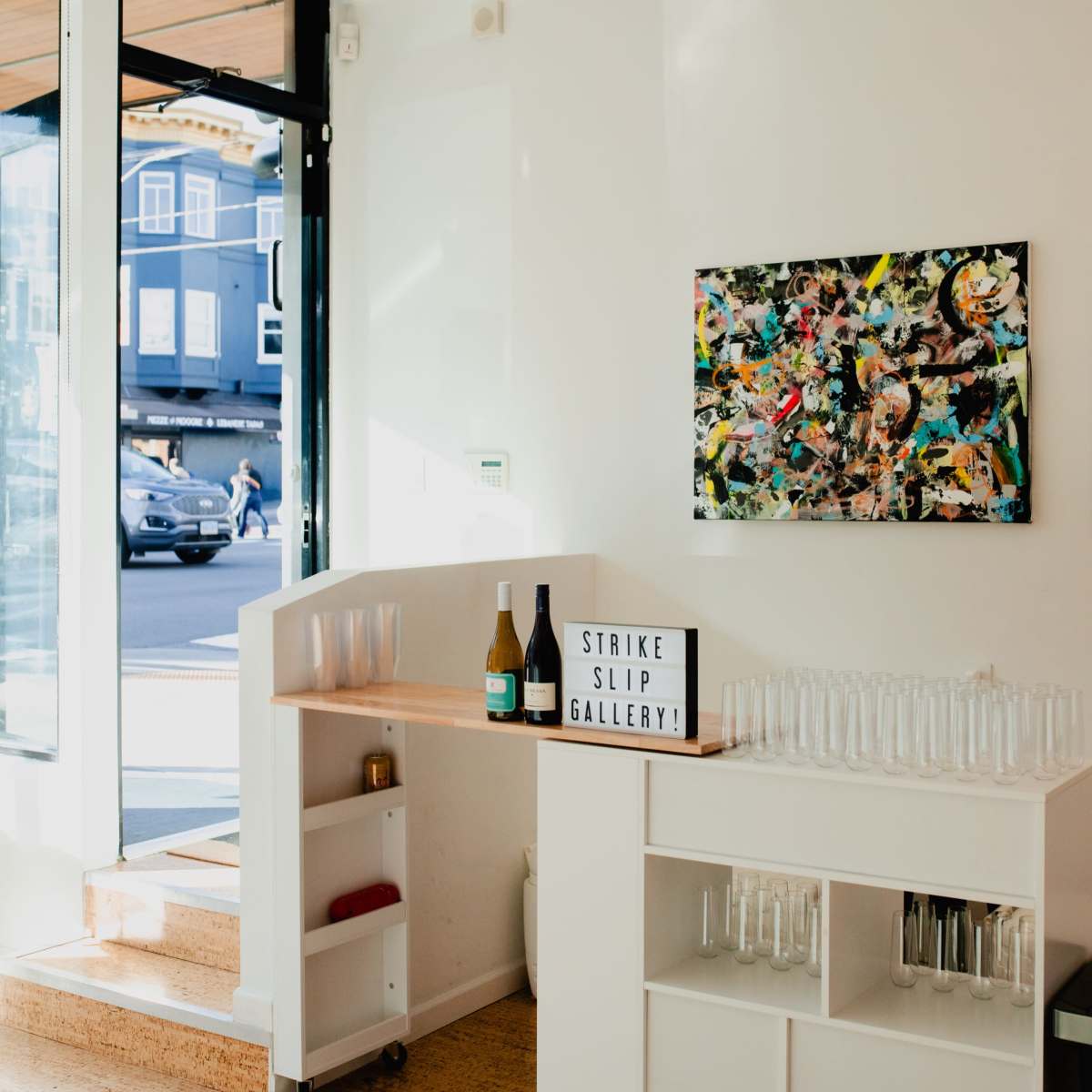 The interior of a bright, modern gallery space featuring a bar area with bottles and glasses, alongside a colorful abstract painting on the wall.