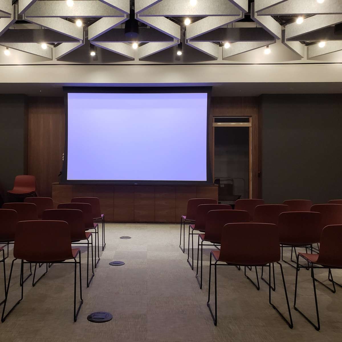 A conference room is set with rows of red chairs facing a blank projection screen.