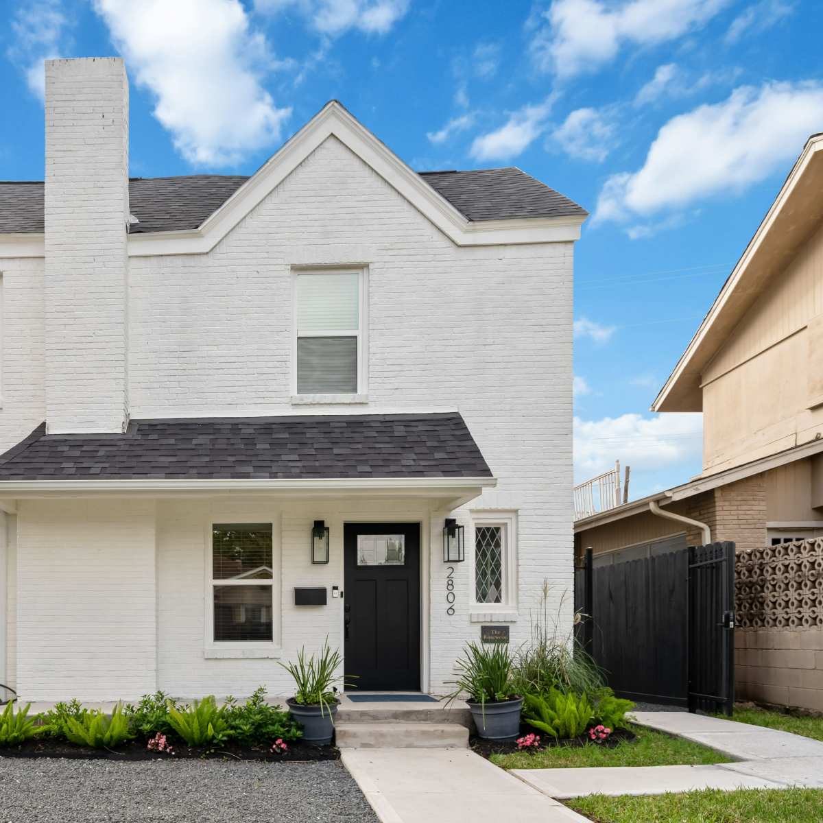 A two-story white brick house with a black roof, surrounded by a landscaped yard featuring green plants and gravel pathways.