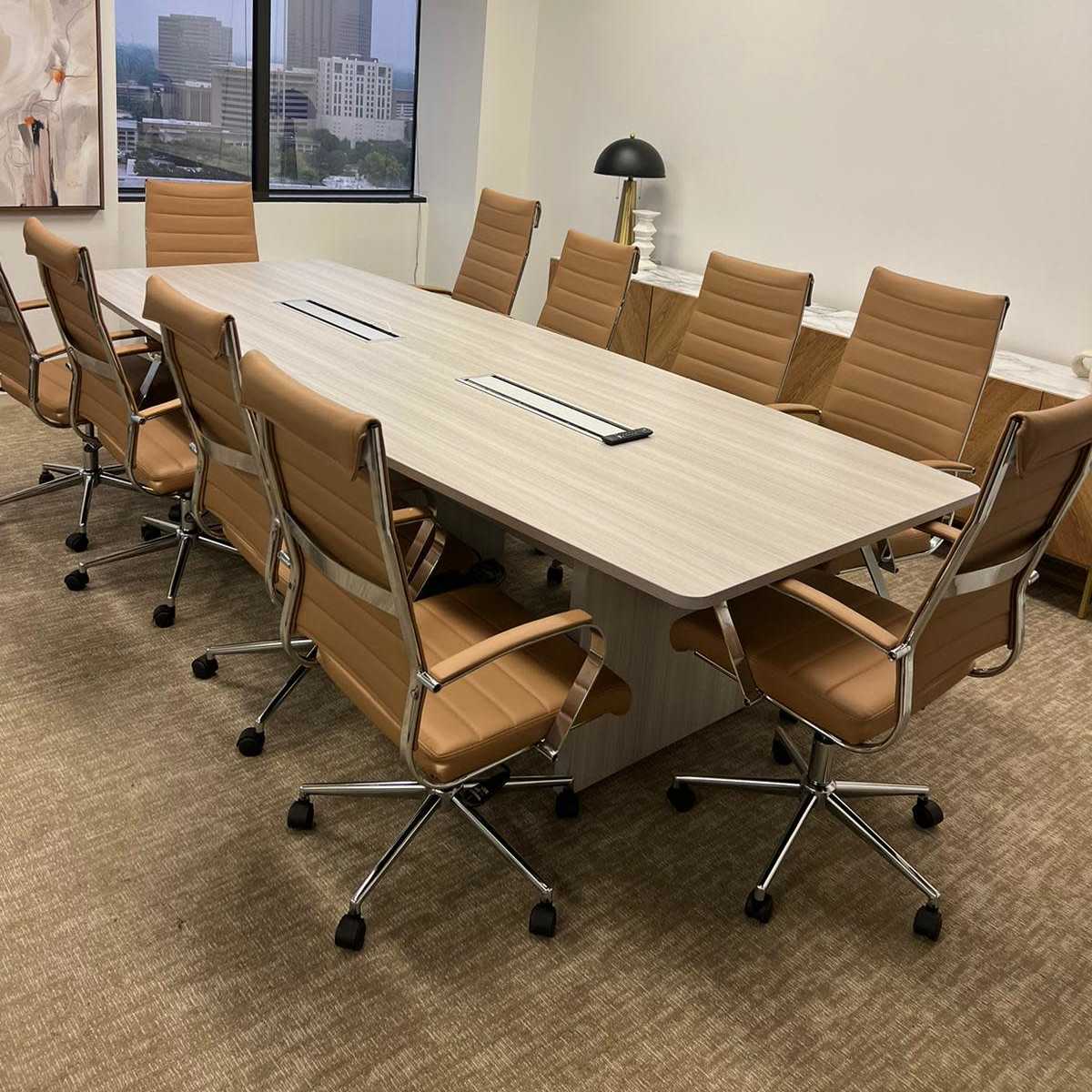 A modern conference room with a long wooden table surrounded by eight beige rolling chairs, and large windows providing a view of a city skyline.