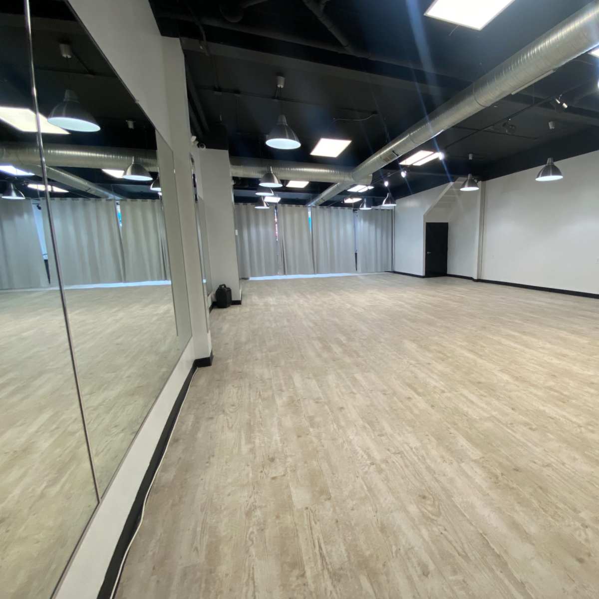An empty studio room with a wooden floor, large mirrors on one wall, and overhead lighting.
