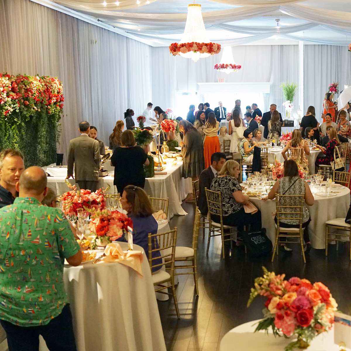 A spacious event hall is filled with guests mingling around tables adorned with flowers and set for a formal occasion.
