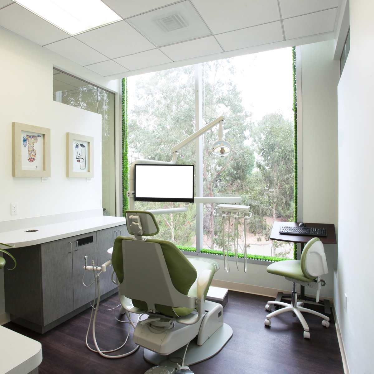 A dental office features a treatment chair and equipment positioned near a large window with greenery visible outside.
