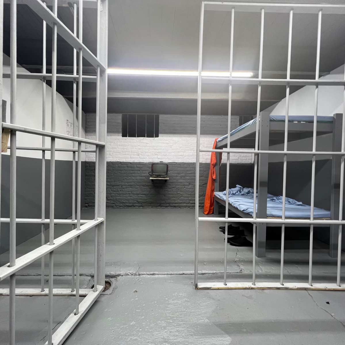An empty prison cell with two barred doors, a bed with blue bedding, and a small television set against a wall.