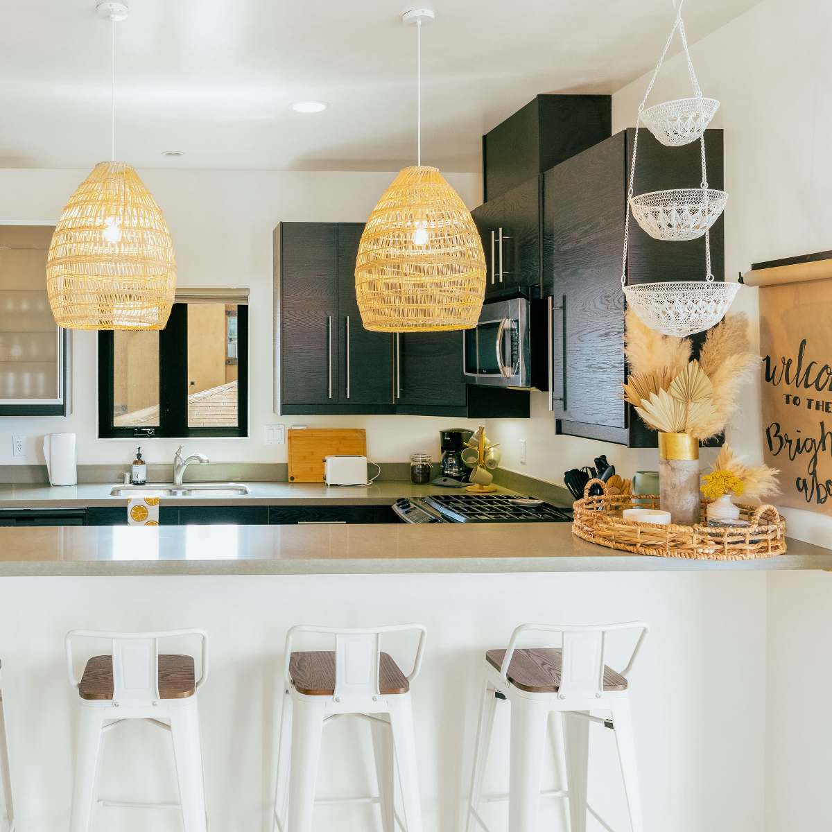 A modern kitchen with a breakfast bar, featuring wooden pendant lights, dark cabinetry, stainless steel appliances, and a white countertop.