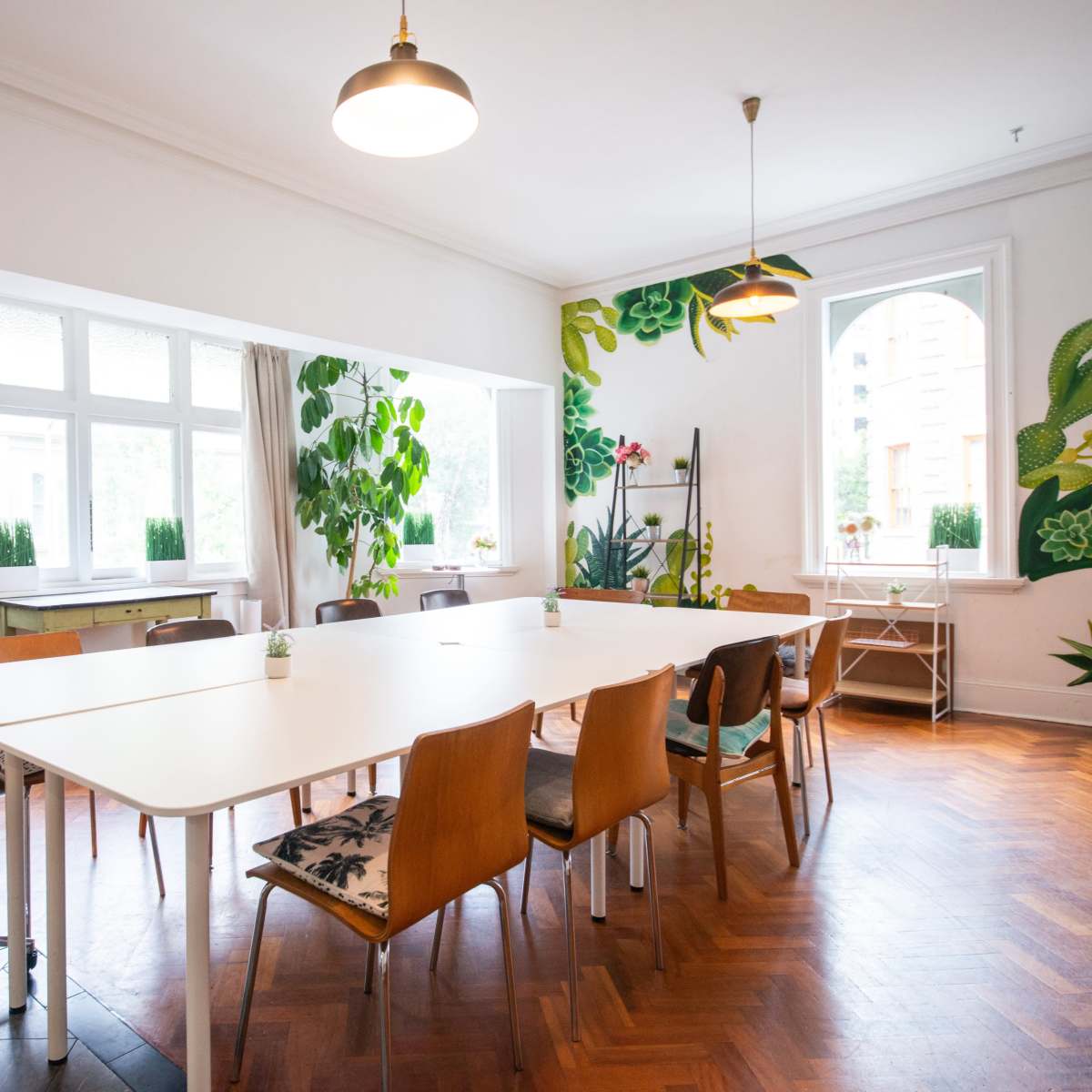 A spacious meeting room features a long white table surrounded by chairs, with large windows and plant-themed wall decorations.