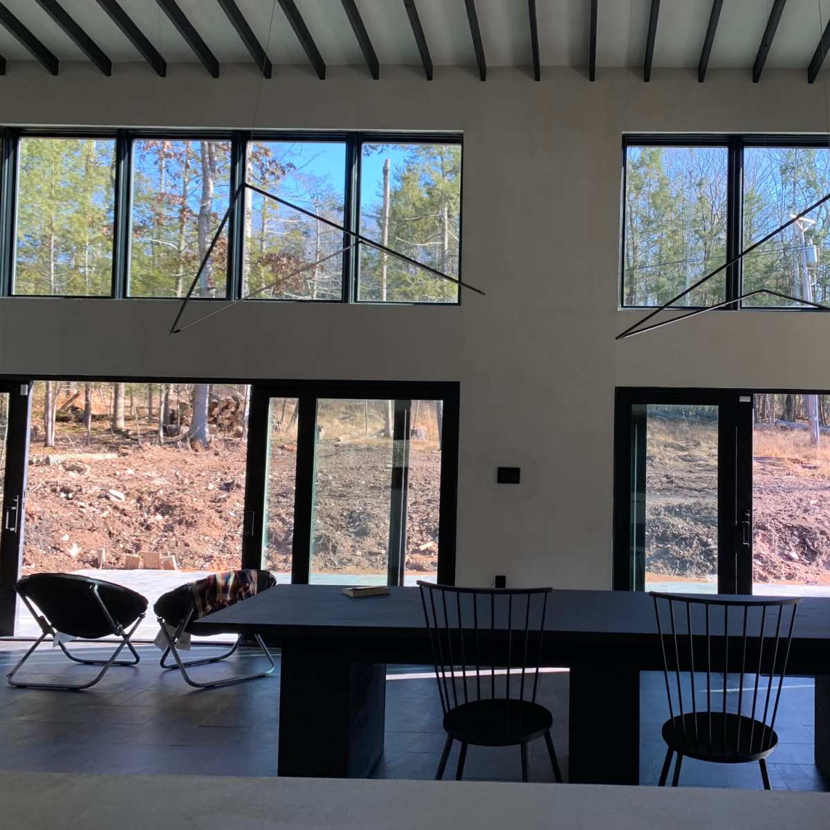 A modern interior space with large windows, a long dark dining table, black chairs, and a view of a wooded area outside.