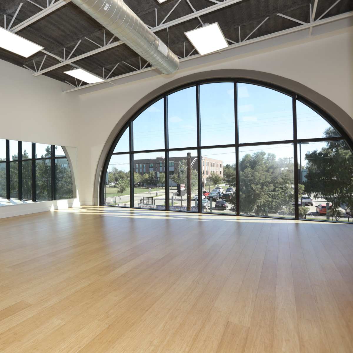 A large, well-lit dance studio features a curved window, a mirror along one wall, and a bamboo floor.