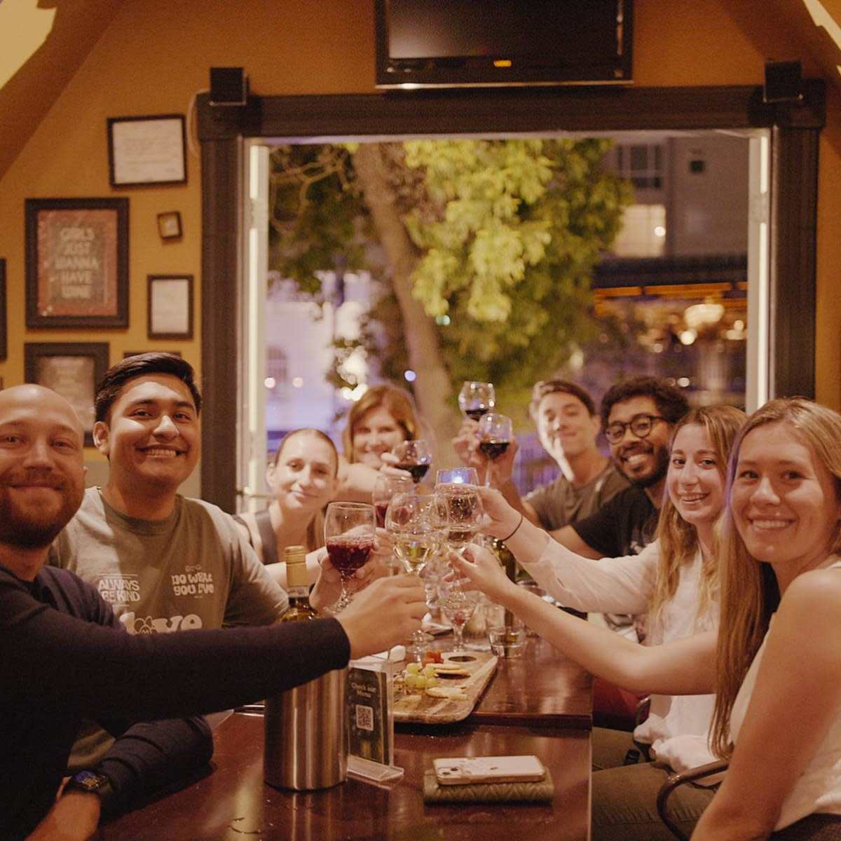 A group of people sit around a table in a warmly lit restaurant, smiling and raising their glasses in a toast.