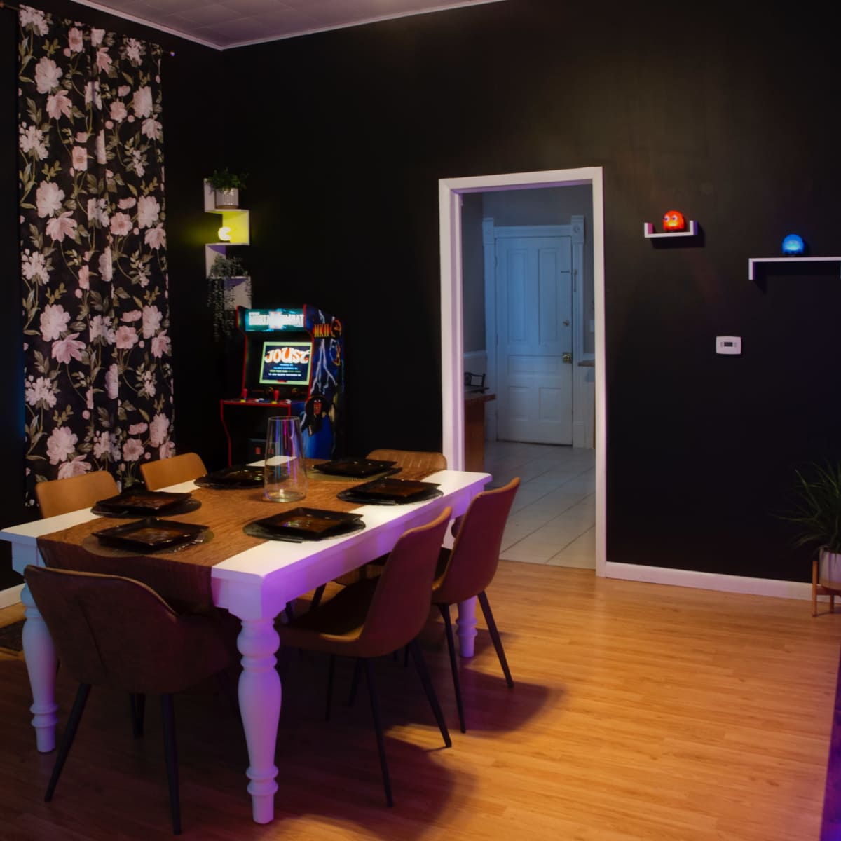 A dining room with a white table set for six, surrounded by wooden chairs, against a wall decorated with floral wallpaper and an arcade game machine in the background.