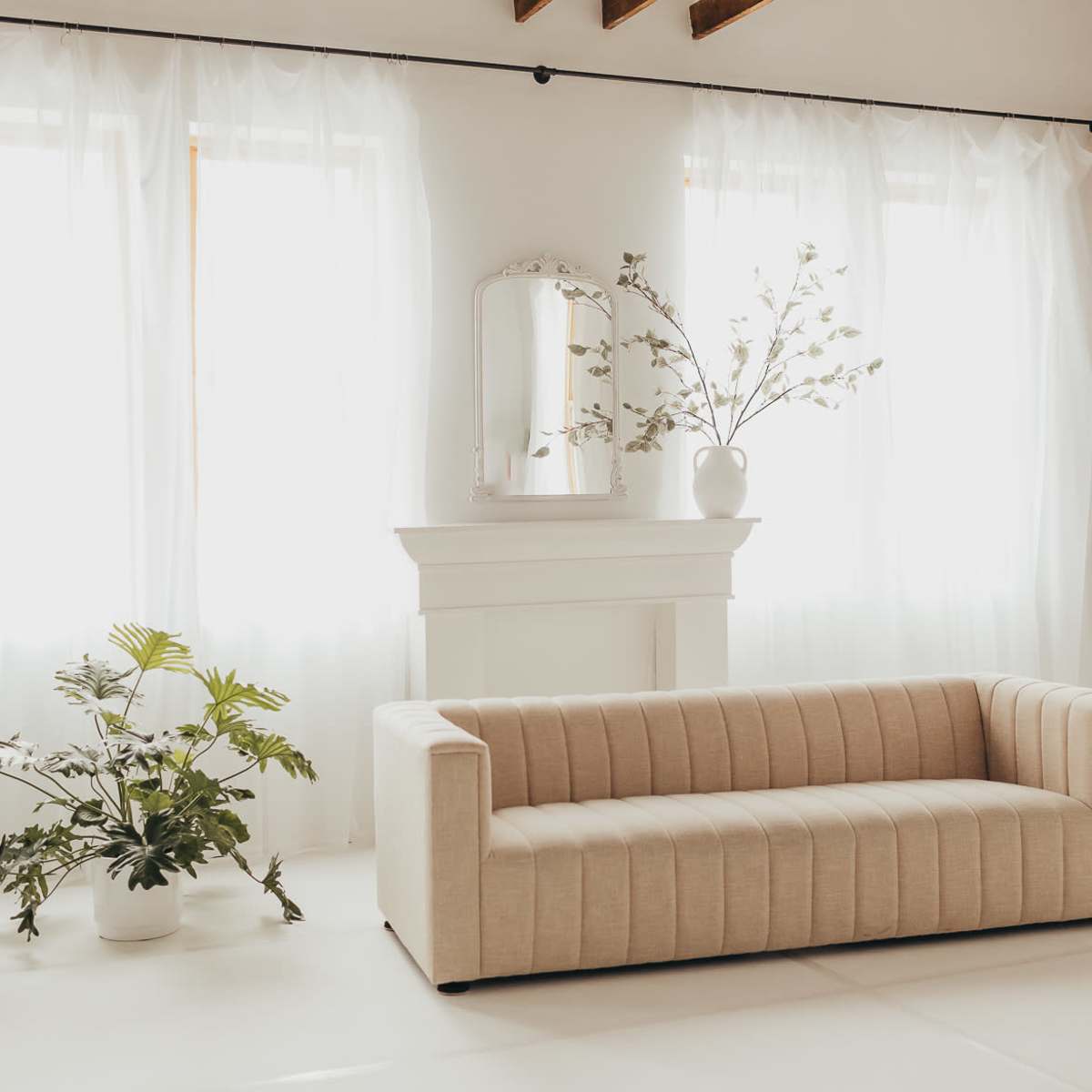 A beige, tufted sofa sits in a bright room with large windows draped in sheer curtains, beside potted plants and a decorative mirror on a white mantel.