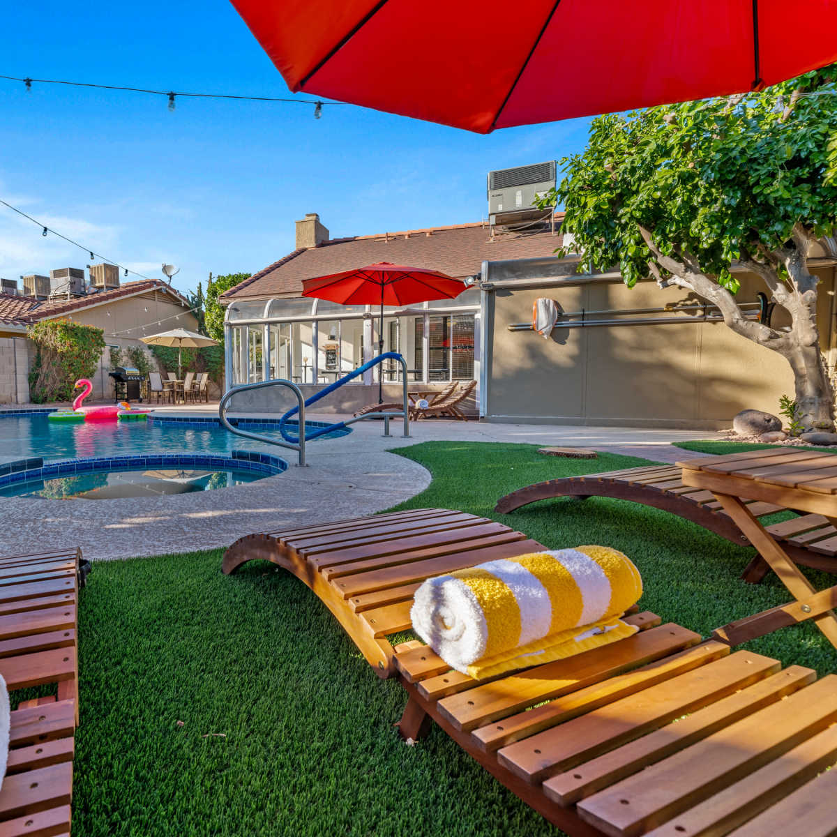 A backyard features a pool surrounded by lounge chairs, an umbrella, and a pink inflatable flamingo.