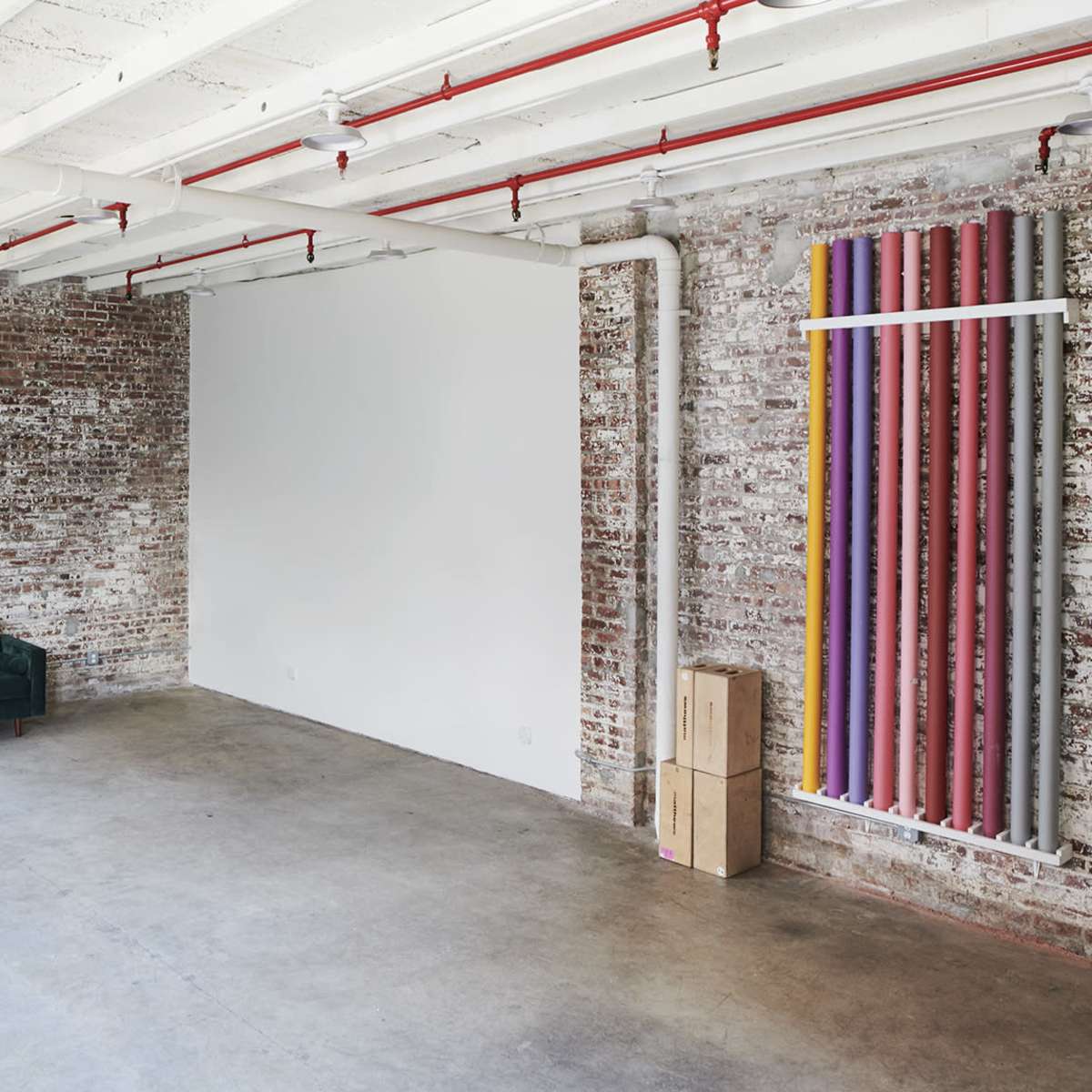 An empty industrial-style room with exposed brick walls, polished concrete floors, a green sofa, and colorful vertical pipes on one side.