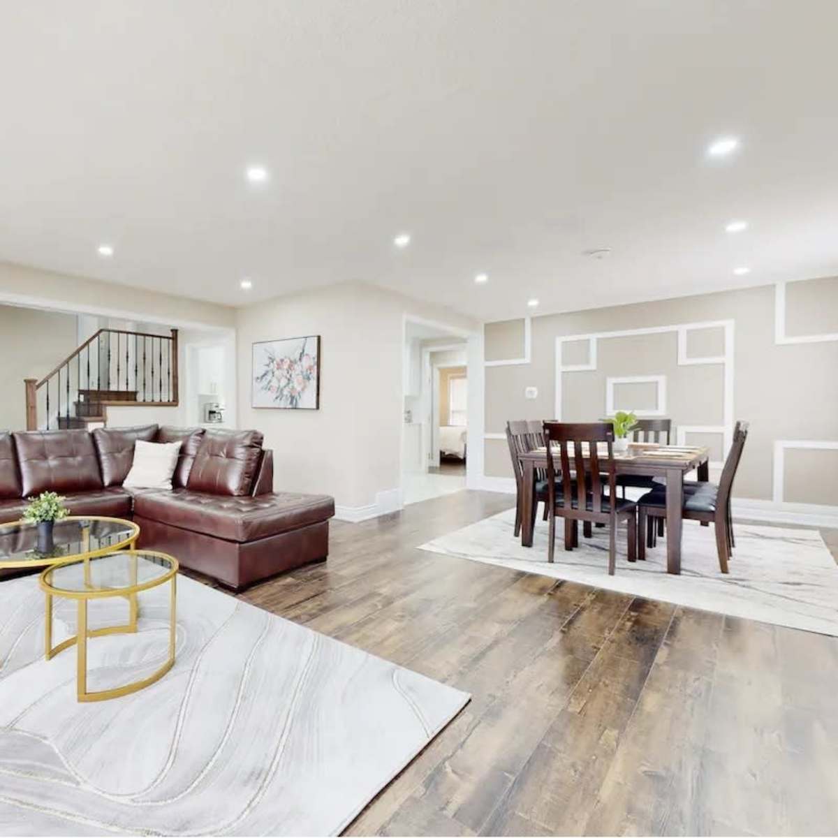 The living room features a brown leather sectional sofa, a glass-top coffee table, and a dining area with a wooden table and chairs, all set on hardwood floors.
