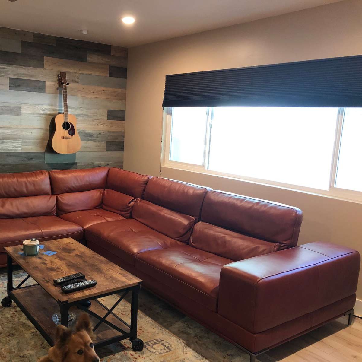 A leather sectional couch is positioned in a living room with a wooden accent wall, an acoustic guitar hanging above, and a coffee table in front.