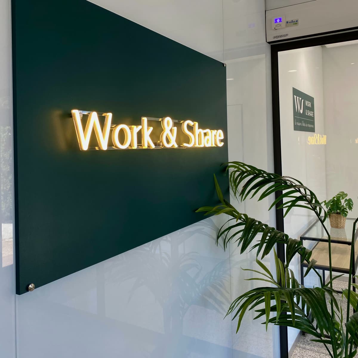 A well-lit sign that reads "Work & Share" mounted on a green wall in an office space, with a plant visible in the foreground.