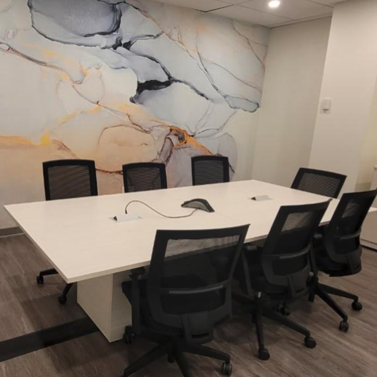 A modern conference room features a large white table surrounded by black chairs, with a mural of abstract shapes on the wall behind it.