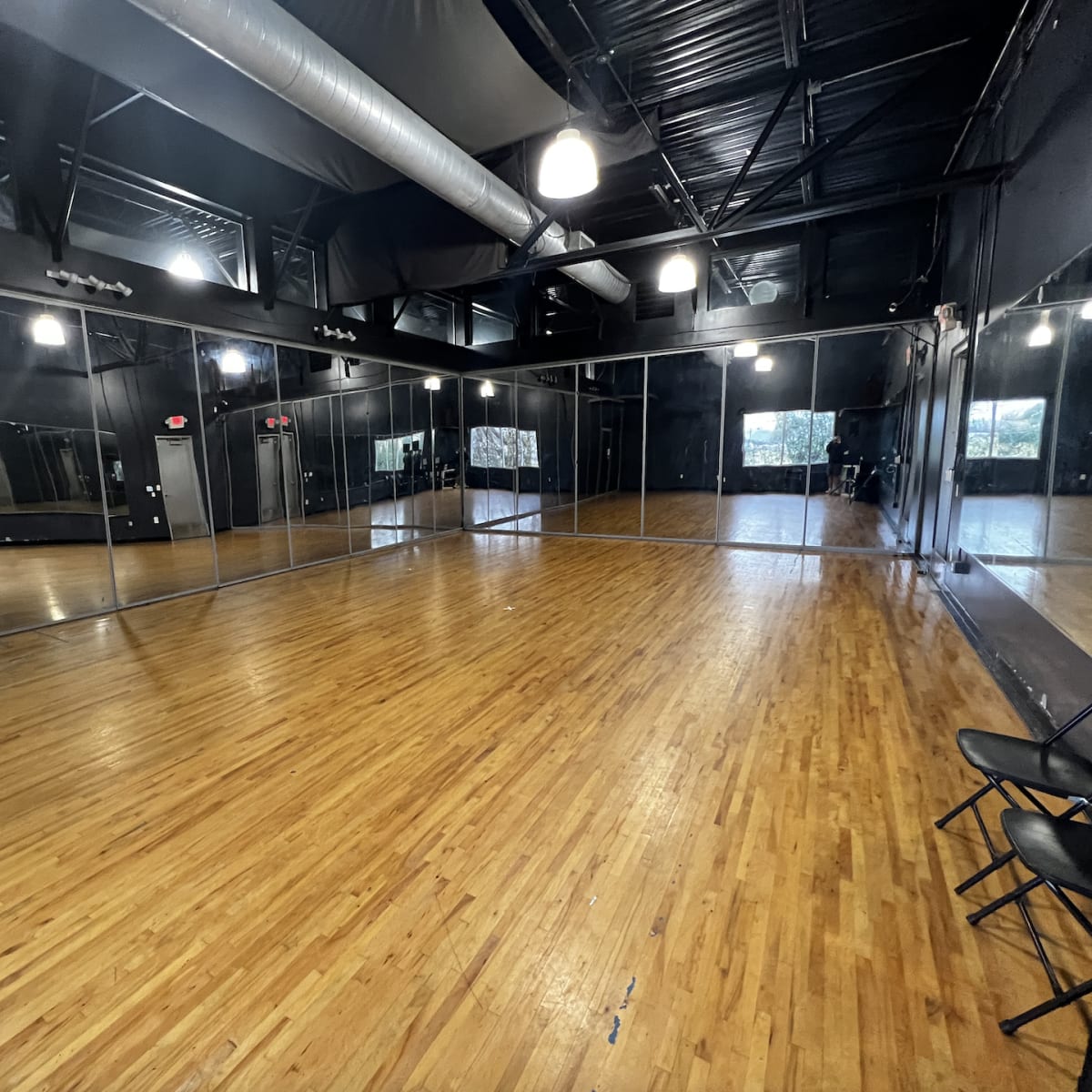 A spacious dance studio featuring wooden flooring, mirrored walls, and several folding chairs against one side.