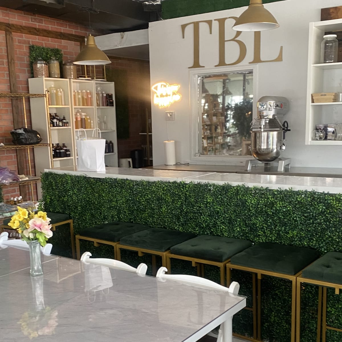 A small cafe interior with a marble-topped table, green upholstered bar stools, and a counter adorned with greenery and shelves of various products.
