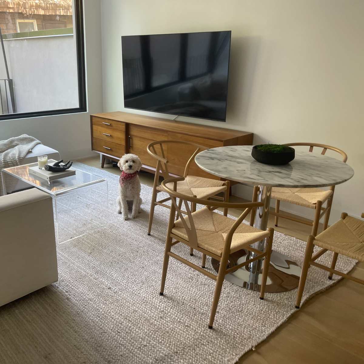 A living room features a sofa, a television on a wooden cabinet, a round marble dining table with chairs, and a dog sitting on a woven rug.