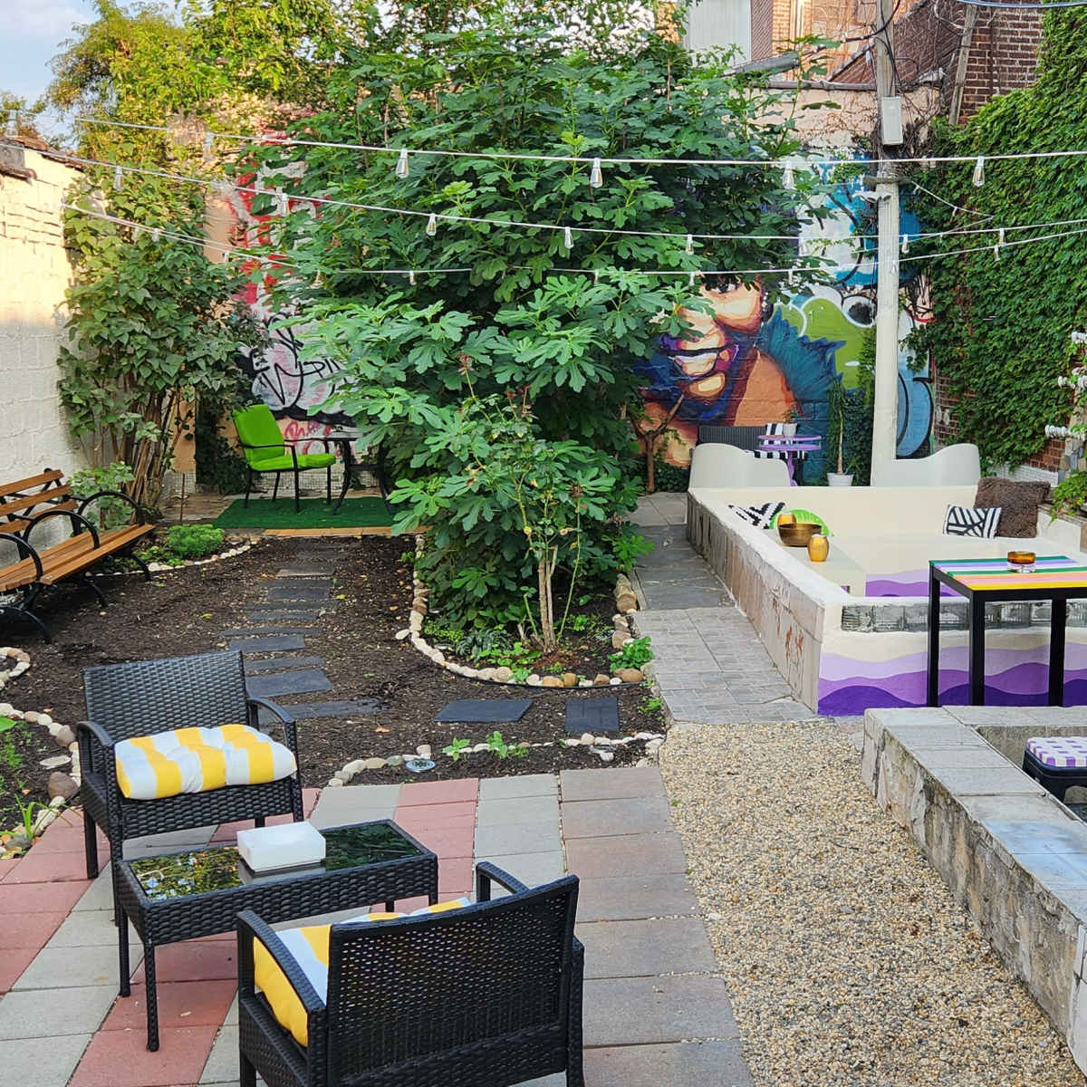 A landscaped backyard with seating areas, a fire pit, and a mural-covered wall, surrounded by greenery.