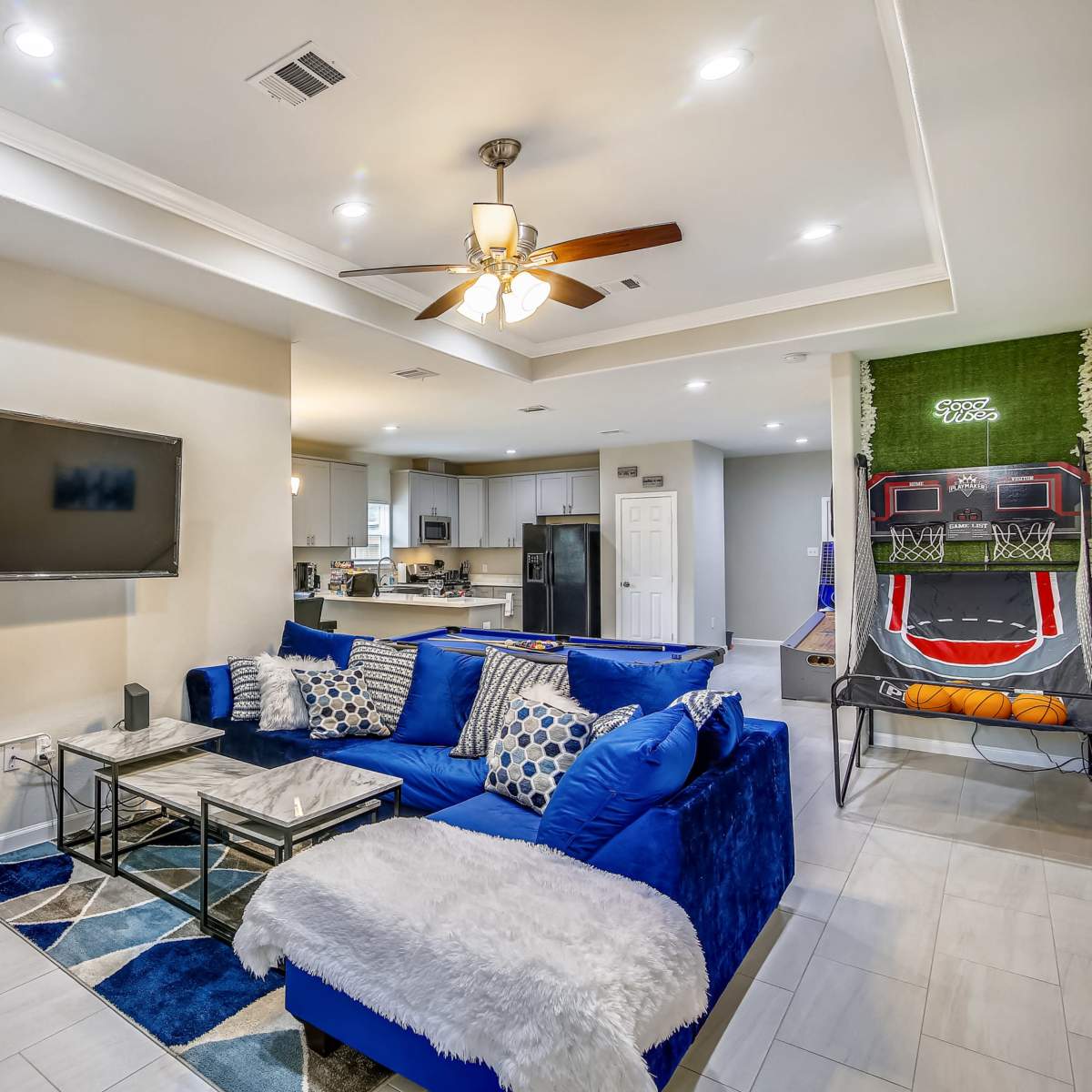 A modern living room featuring a blue sectional sofa, a flat-screen TV mounted on the wall, and a kitchen visible in the background, along with a basketball shooting game in the corner.