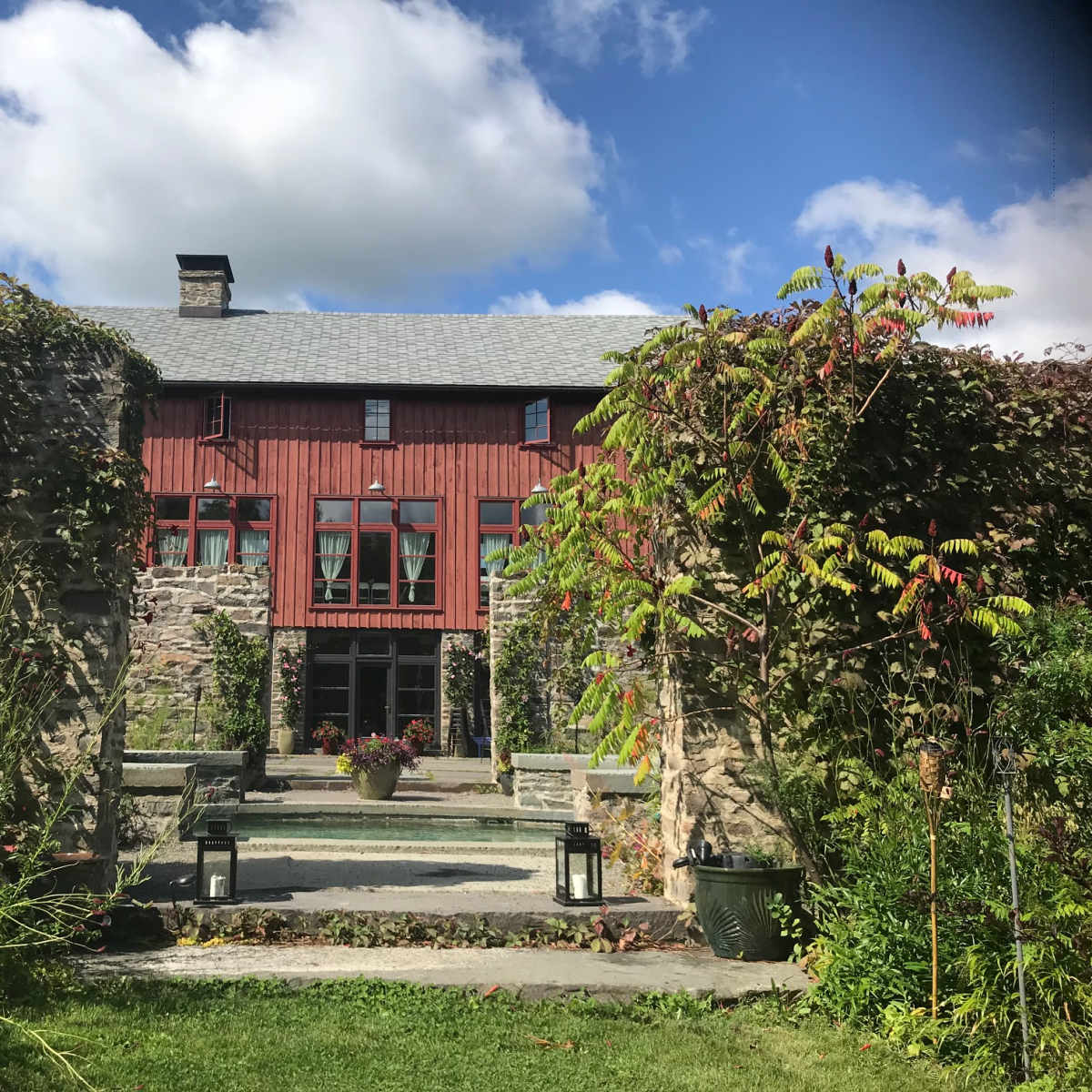 A red and stone house is framed by lush greenery and a pathway leading to its entrance under a blue sky with clouds.