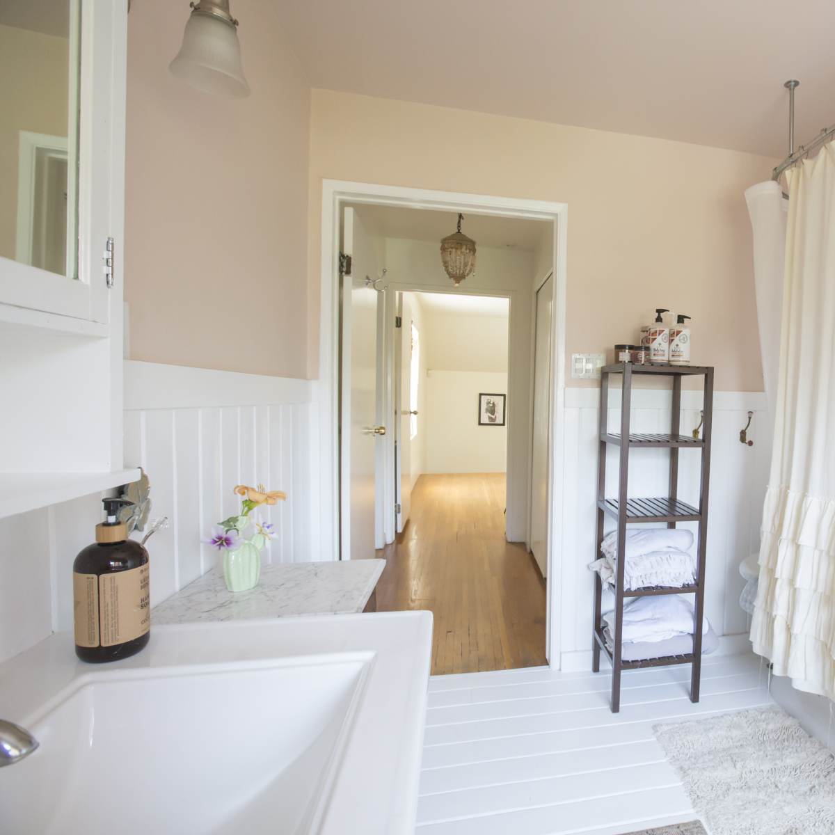 A bright bathroom with a white shower curtain, a marble countertop with a soap dispenser, and a shelf holding folded towels.