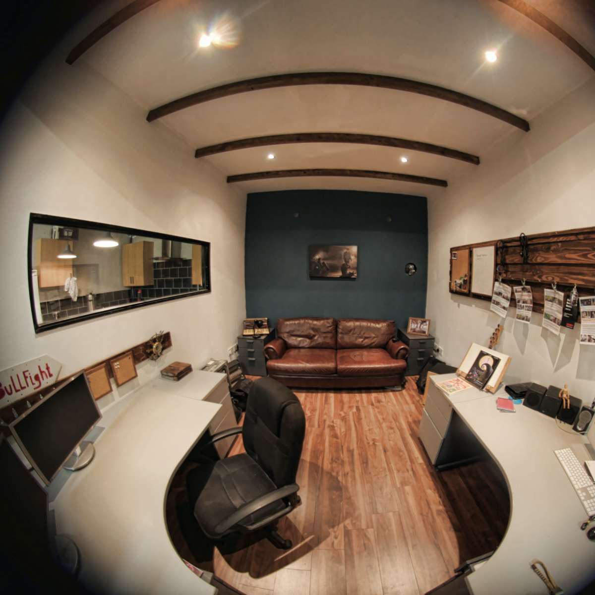 A small office space featuring a brown leather sofa, two workstations with computers, and wooden beams on the ceiling.