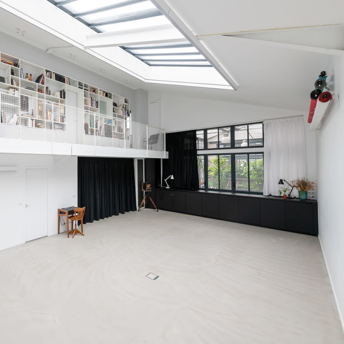 A spacious, modern interior with a high ceiling, a loft area lined with bookshelves, and large windows allowing natural light to enter.