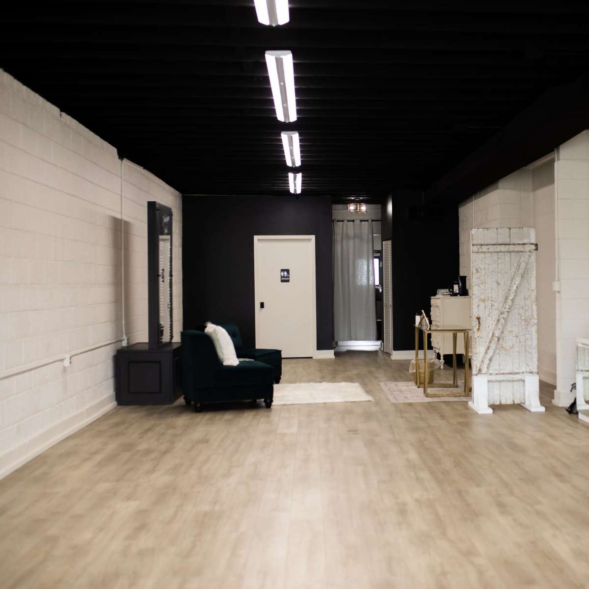 A spacious interior with light-colored walls, a wooden floor, and various furniture pieces along the sides, including a green chair, a small table, and a large mirror.